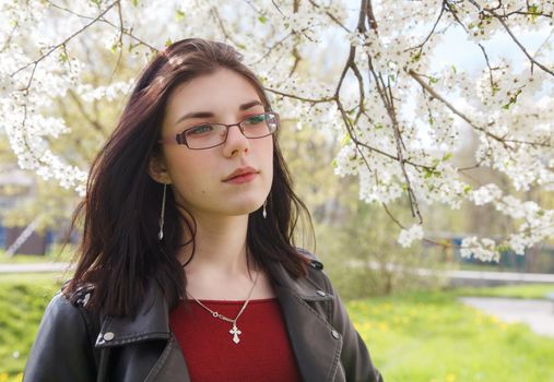 portrait of young beautiful brunette girl in black jacket and burgundy sweater standing near cherry blossoms on sunny spring day. outdoor closeup