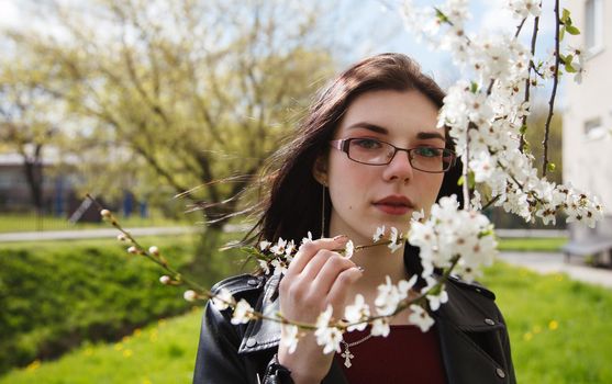 portrait of young beautiful brunette girl in black jacket and burgundy sweater standing near cherry blossoms on sunny spring day. outdoor closeup