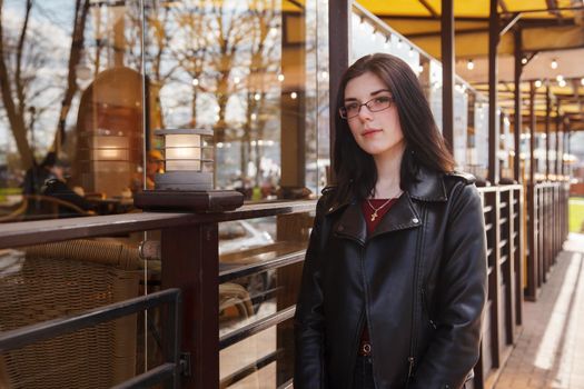 young beautiful brunette girl in black jacket and jeans  stands near a cafe on a city street on summer day