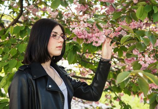 portrait of young beautiful brunette girl in black jacket standing near cherry blossoms on sunny spring day. outdoor closeup