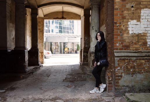 sad young girl in black jacket and jeans stands in old arch on city street on summer day