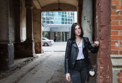 sad young girl in black jacket and jeans stands in old arch on city street on summer day. closeup outdoor