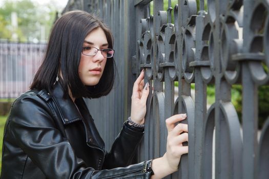 sad young girl in black jacket stands near metal fence on city street on summer day. closeup outdoor