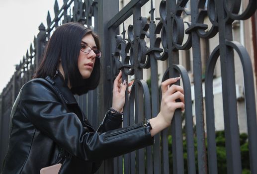 sad young girl in black jacket stands near metal fence on city street on summer day. closeup outdoor