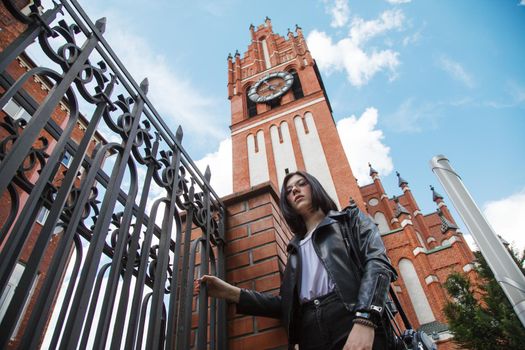 young beautiful brunette girl in black jacket and jeans stands near gothic church on summer day