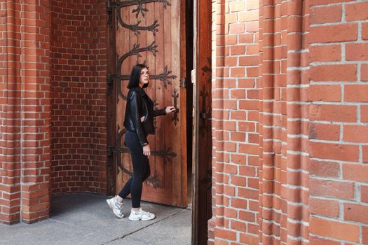 young beautiful brunette girl in black jacket and jeans stands near door of gothic church on summer day