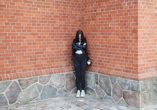 young beautiful brunette girl in black jacket and jeans stands near wall of gothic church on summer day