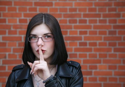 young beautiful brunette girl in black jacket shows a gesture quietly near brick wall. outdoor closeup