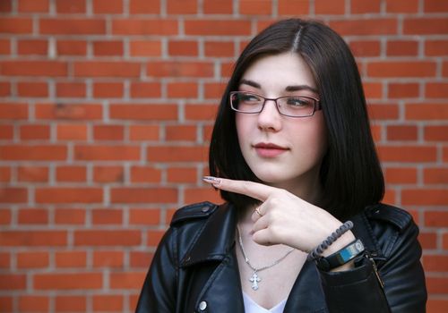 positive beautiful brunette girl in black jacket points to the side. presenting your product. outdoor closeup on brick wall background