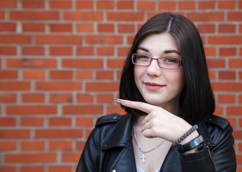 positive beautiful brunette girl in black jacket points to the side. presenting your product. outdoor closeup on brick wall background