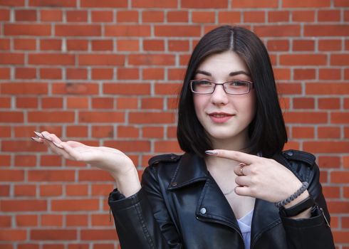 positive beautiful brunette girl in black jacket points to the side. presenting your product. outdoor closeup on brick wall background