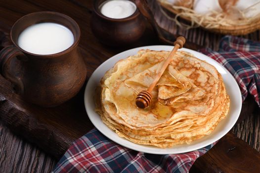 Homemade thin pancakes with honey stacked in a stack, on a wooden table with a mug of milk, a pot of sour cream and eggs in a basket. Traditional Slavonian, pagan holiday (Maslinitsa). Country style food.