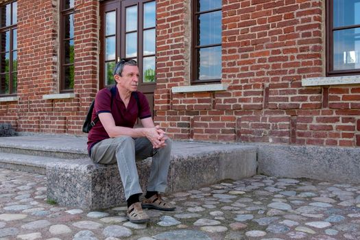 Close-up of an adult pensioner in summer clothes and with a backpack sits and rests against the backdrop of a red brick building. Life style. Selective focus.