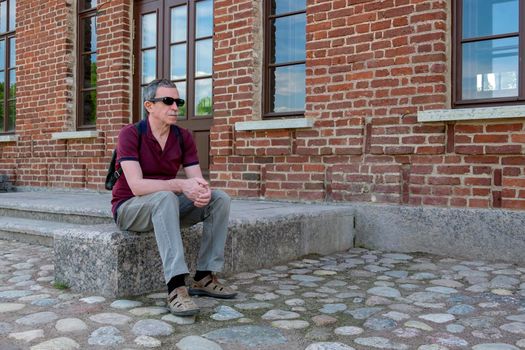 Close-up of an adult pensioner in summer clothes and with a backpack sits and rests against the backdrop of a red brick building. Life style. Selective focus.