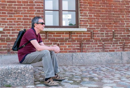 Close-up of an adult pensioner in summer clothes and with a backpack sits and rests against the backdrop of a red brick building. Life style. Selective focus.