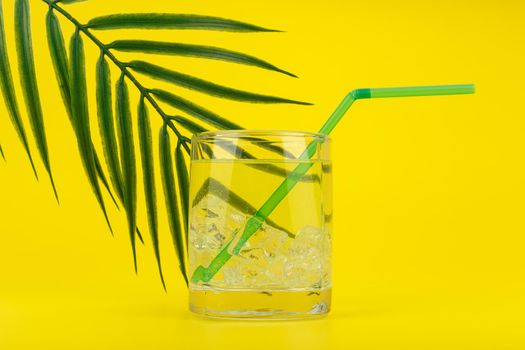 Summer composition with transparent glass with soda, lemonade or cocktail filled with ice cubes on yellow background decorated with palm leaf. Concept of cold and refreshing summer drinks