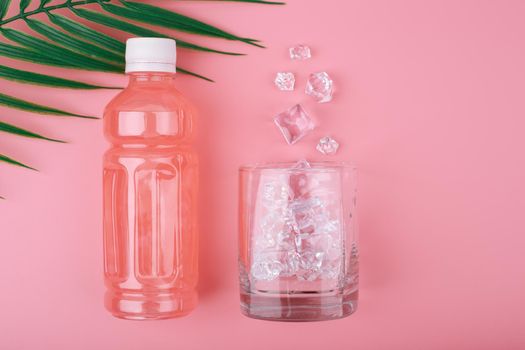 Creative flat lay with pink lemonade in a bottle and glass with spilled ice on pink background with palm leaf. Concept of refreshing cold drinks for summer or detox drinks
