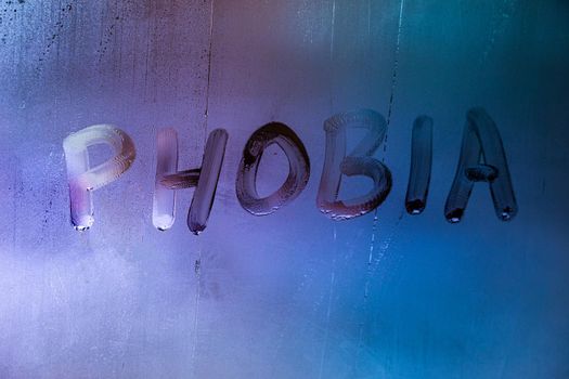 the word phobia handwritten on wet foggy window glass surface with blue background light.