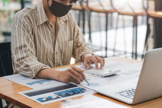 Businessman or accountant wear mask working on desk office with using a calculator to calculate the numbers, finance accounting concept.