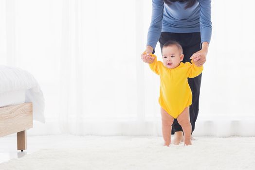 Asian little baby girl taking first steps learning to walk with mom help support the cute unstable walking toddler at home in bedroom. Happy family first steps parenthood concept