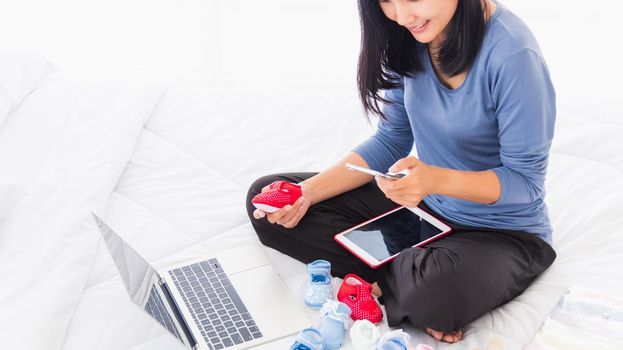 Asian mother shopping online for her baby clothes on laptop computer and tablet on the bed she makes purchase new baby clothes for an unborn baby preparing for the new child