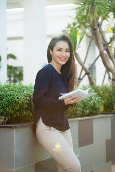 Portrait of smiling female student reading book while standing outdoor on terrace of campus cafe in sunny day. Education, lifestyle and people concept.