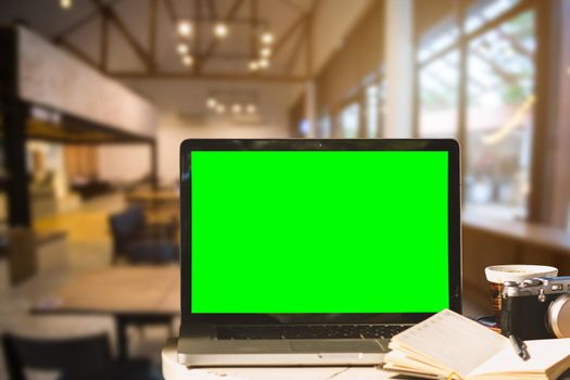 Mockup image of laptop with blank green screen with camera,notebook,coffee cup on wooden table of In the coffee shop background.