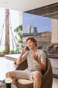 Asian man talking on a phone while sitting outdoors in sunny summer day.