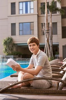 Young asian man reading book by the pool on a sunny summer day