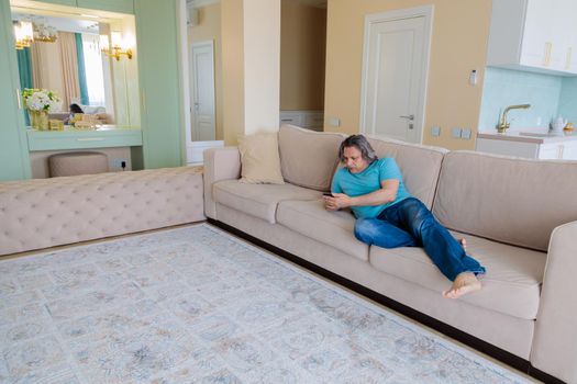 An adult man browses social networks in a mobile phone while lying on a sofa in an apartment.