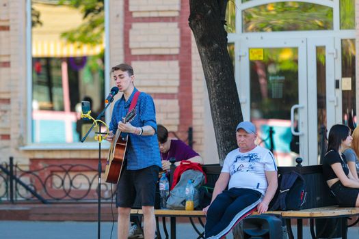 A young guy stands with a guitar on a central street, plays and sings, earning money.
