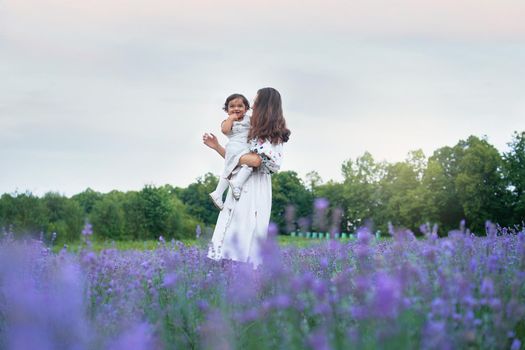Young woman posing with baby girl on hands in summer lavender field. Side view of mother wearing dress carrying little laughing lovely kid. Concept of nature beauty, family.