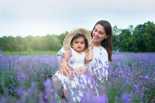 Young happy mother sitting between purple flowers with kid wearing straw hat and looking at camera. Smiling woman posing with baby daughter on knees in lavender field. Concept of nature, motherhood.