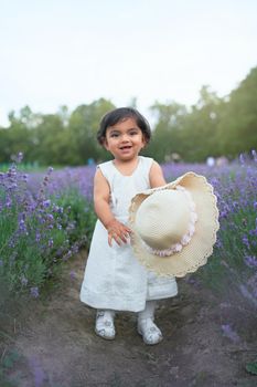 Front view of adorable young female kid posing with straw hat in blooming aromatic lavender field and laughing. Little pretty baby girl wearing lovely white dress enjoying time outdoors.