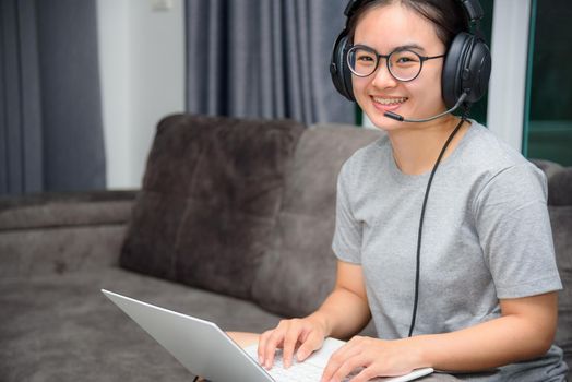 Asian young woman student with headphones sitting on the sofa smile looking up happy study online class college learning internet education, Teenage girl work distance on a laptop computer from home