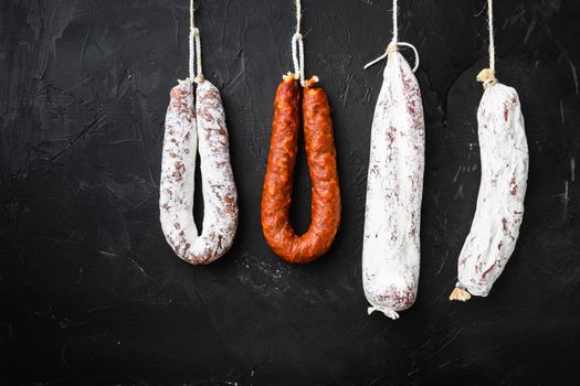 Spanish salami, fuet and salchichon sausages hang from a rack on black surface.