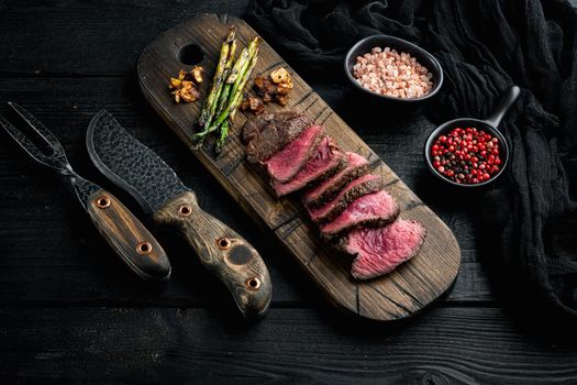 Grilled fillet mignon beef steak set, with onion and asparagus, on wooden serving board, with meat knife and fork, on black wooden table background