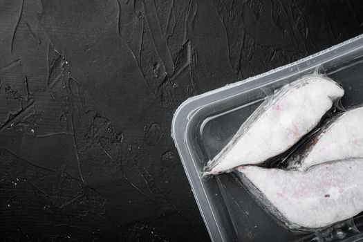 Frozen Greenland halibut steaks vacuum pack set, on black dark stone table background, top view flat lay, with copy space for text