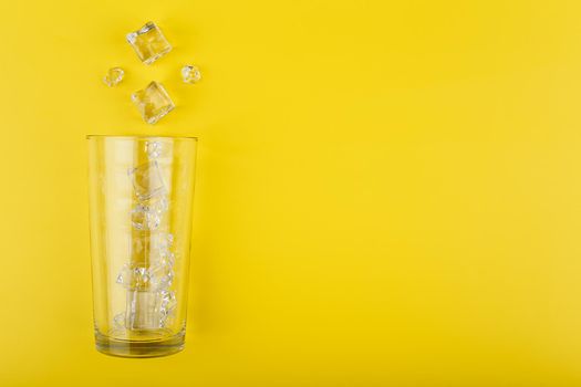 Creative flat lay with empty transparent glass with spilled ice cubes on yellow background with copy space. Concept of refreshing summer drinks and cocktails