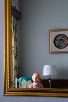 Creepy looking plastic toy doll reflection in gold framed mirror household scene with woven wall art and floral curtains, South Africa