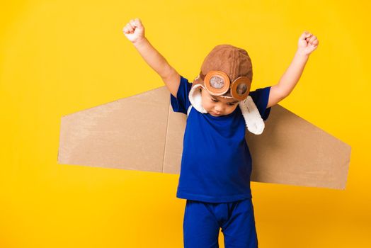 Happy Asian handsome funny child or kid little boy smile wear pilot hat play and goggles raise hand up with toy cardboard airplane wings flying, studio shot isolated yellow background, Startup freedom