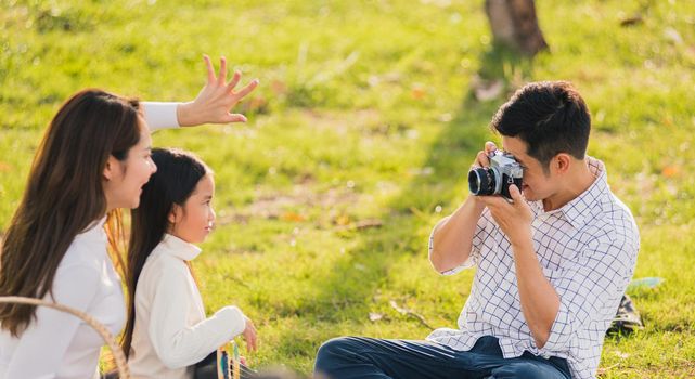 Happy Asian young family father, mother and child having fun and enjoying outdoor together sitting on the grass party with shooting photos by retro camera a picnic in the garden park on a sunny day