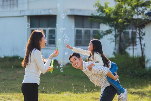 Happy Asian lifestyle family mother, father and little cute girl child having fun together and enjoying outdoor play blowing soap bubbles in the garden park on a sunny day, summertime