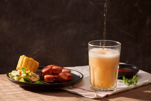 Pouring beer into glass with sausage, ketchup, corn and salad on wooden table