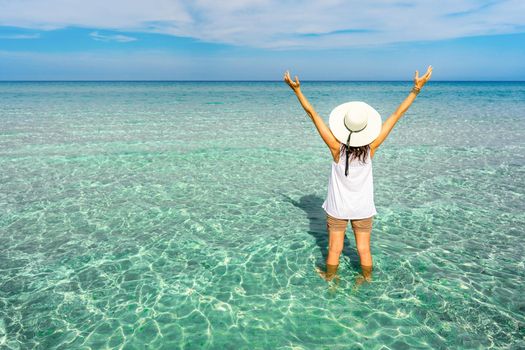 View from back of hipster unrecognizable millennial happy young woman with a large white hat in vacation resort on a crystal clear tropical sea water open arms up to the blue sky looking to horizon