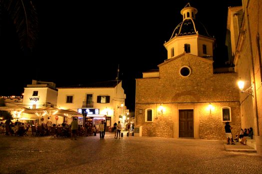 Altea, Alicante, Spain- June 12, 2021: People having drinks and enjoying nice weather in terraces in the main square of Altea village in Spain. The Virgin of the Consol church at night.
