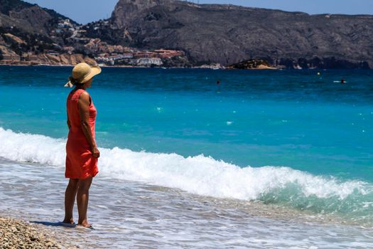 Altea, Alicante, Spain- June 12, 2021: Woman in red dress and hat on Cap Negret beach on a sunny day of Spring in Altea, Alicante, Spain