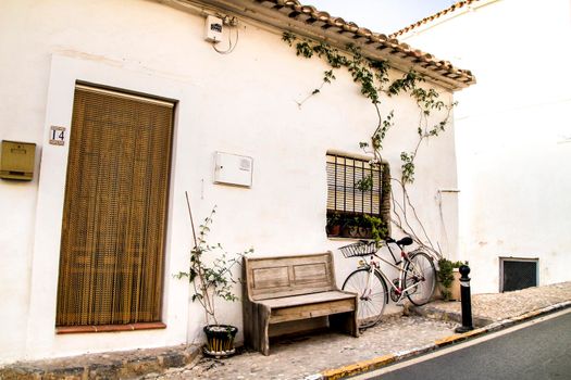 Altea, Alicante, Spain- June 12, 2021: Bicycle resting on the typical whitewashed facade of a house netx to a wooden bench in Altea, Alicante, Spain
