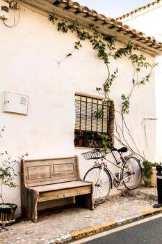 Altea, Alicante, Spain- June 12, 2021: Bicycle resting on the typical whitewashed facade of a house netx to a wooden bench in Altea, Alicante, Spain