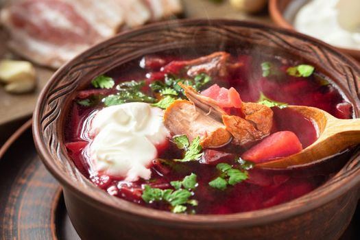 Freshly cooked borscht - traditional dish of Russian and Ukrainian cuisine in earthenware dishes with bacon, bread, sour cream and garlic, close up.
